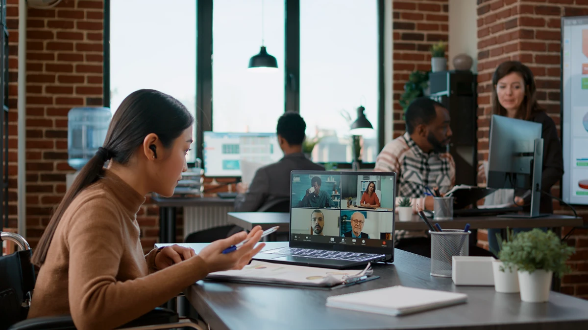 A woman on a video call in a hybrid office while coworkers meet in the background
